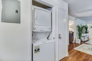 Una lavanderia con lavatrice e asciugatrice a carica frontale, un quadro elettrico e una porta che dà su un corridoio con cucina e sala da pranzo.