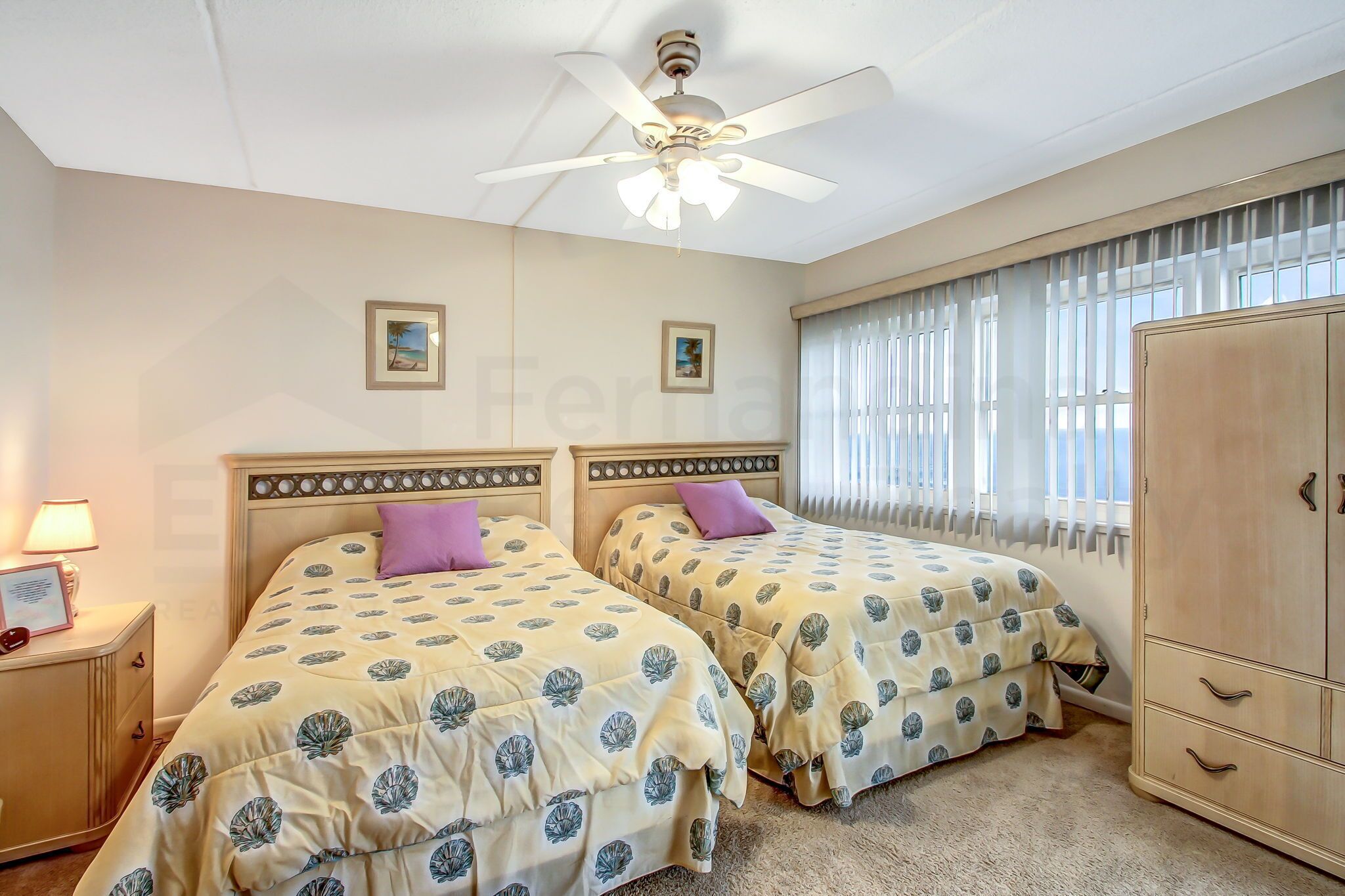 A bedroom with two beds, a ceiling fan, a nightstand with a lamp, and a wardrobe.