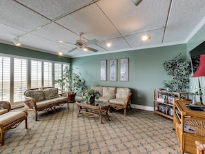 A living room with a ceiling fan, large windows, and a bookshelf. - B7 Amelia South (Fernandina Beach)