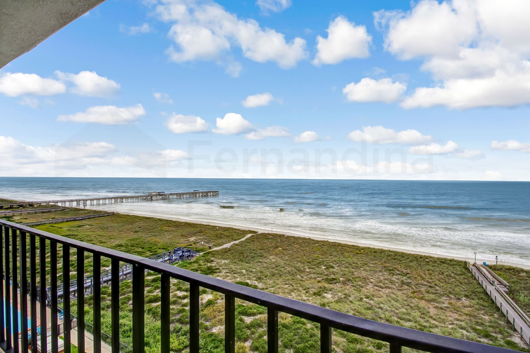 Une vue sur la plage depuis un balcon doté d’une balustrade en bois, une jetée s’étendant dans l’océan et une zone herbeuse au premier plan.
