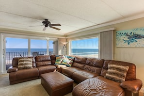 A spacious living room with a brown leather sectional sofa, a ceiling fan, and a large window offering a view of the ocean. - B7 Amelia South (Fernandina Beach)