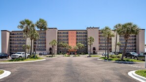 Hotel de varios pisos con entrada central, rodeado de estacionamiento y palmeras.