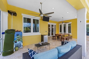 A bright yellow room with a ceiling fan, a TV, a dining area with a table and benches, and a blue sofa. - Sunrise Island Retreat (Fort Pierce)