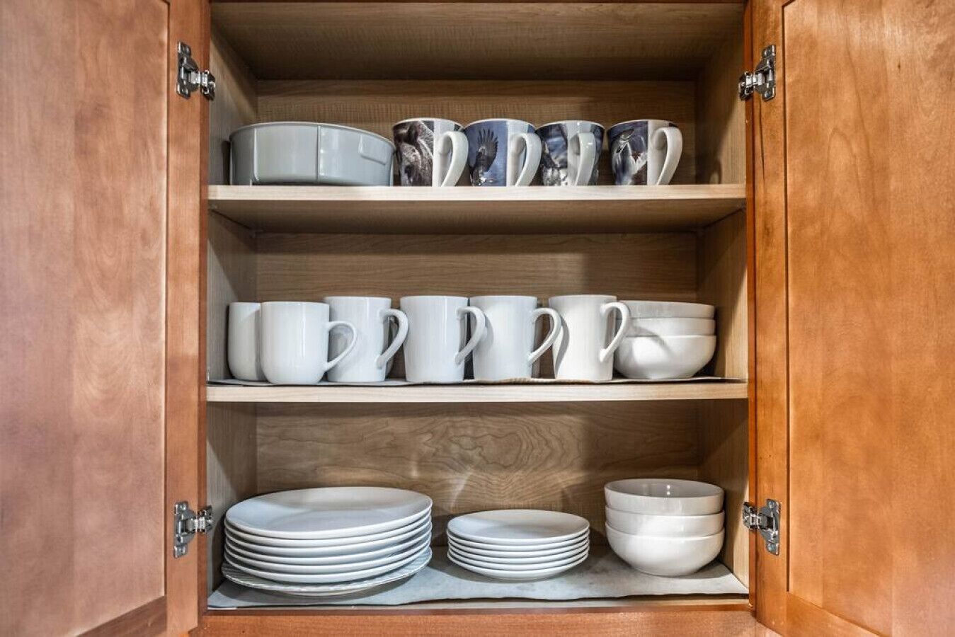 A wooden cabinet with three shelves, each holding white mugs, bowls, and plates.