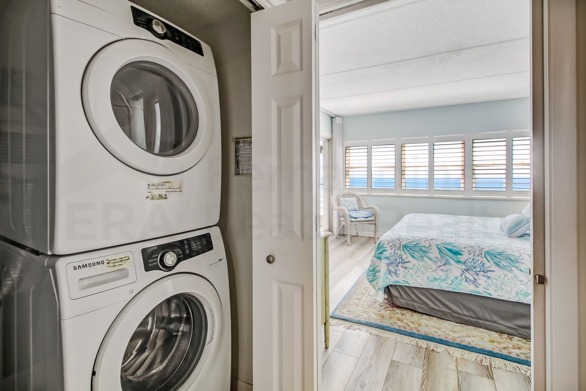 Un espace buanderie avec des lave-linge et des sèche-linge superposés, une chambre avec un lit et une fenêtre, et une porte donnant sur une autre pièce.