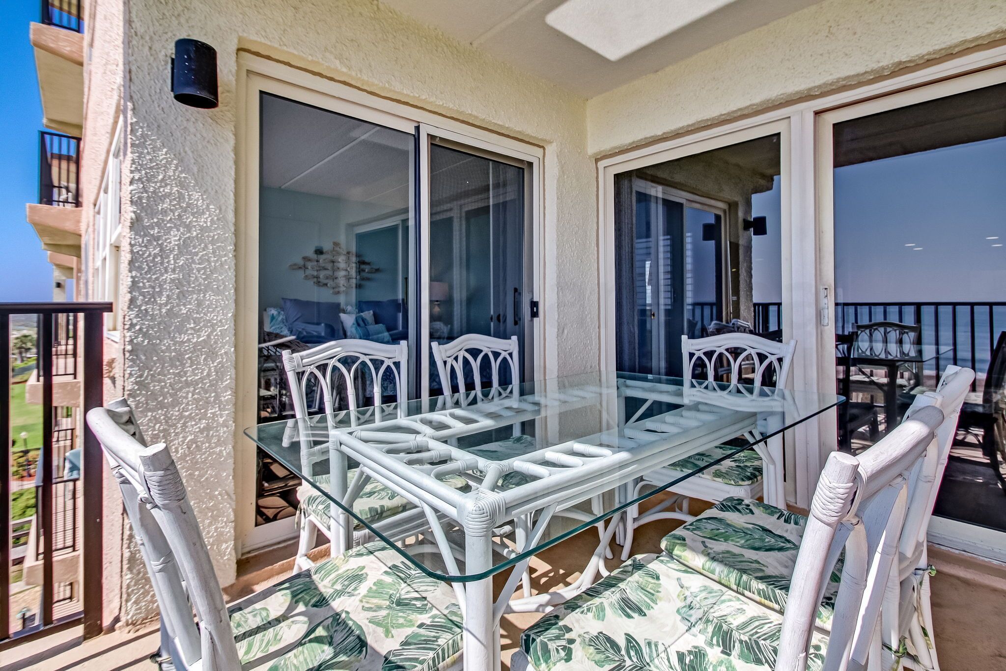 A balcony with a glass table and white chairs, a view of the sea, and a building in the background.