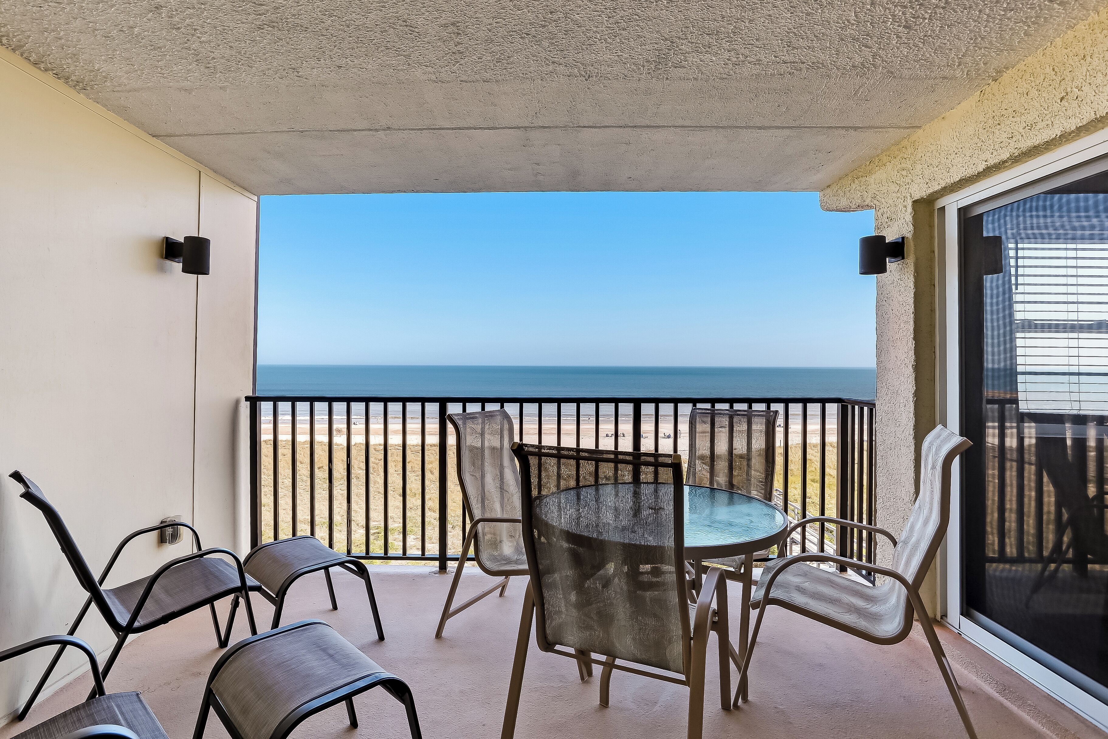 A balcony with a glass table and chairs, overlooking a beach.