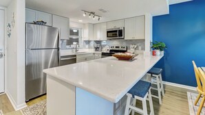 A modern kitchen with white cabinets, a stainless steel refrigerator, a microwave, and a large island with bar stools. - C5 Amelia South (Fernandina Beach)