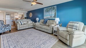 A living room with a beige sofa set, a patterned rug, a ceiling fan, and a painting of palm trees. - C5 Amelia South (Fernandina Beach)