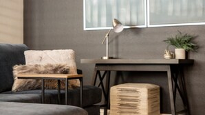 Un salon moderne avec un canapé gris foncé, une petite table d’appoint en bois, une lampe, une plante et une grande fenêtre avec des stores.
