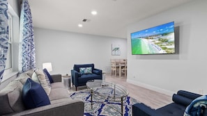 A living room with a sofa, armchair, coffee table, and a TV displaying a beach scene. - Ocean Drive Villas- Unit A (Fort Pierce)