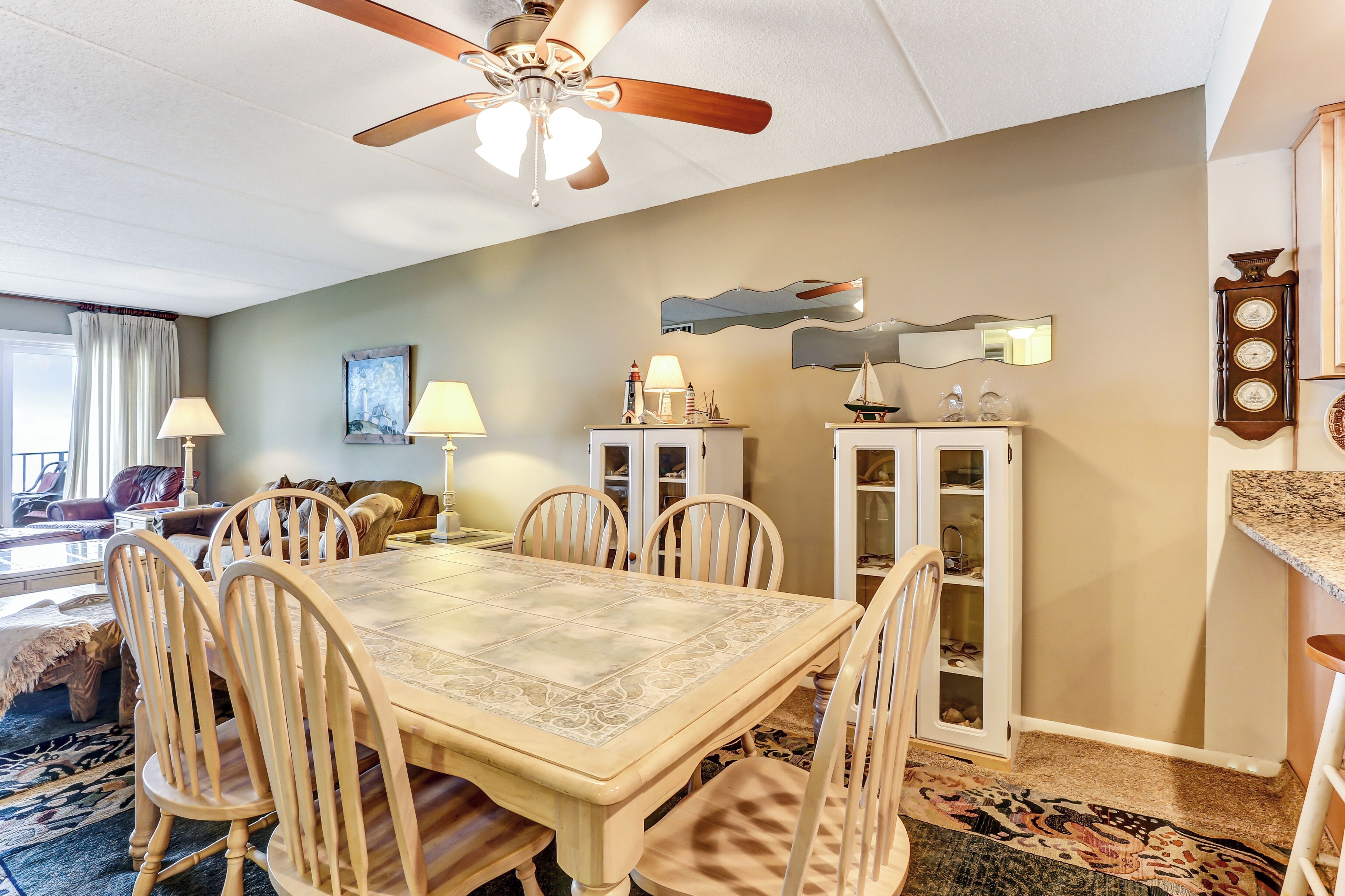 Un coin repas avec une grande table, des chaises et un ventilateur de plafond. On y trouve des armoires avec des portes en verre et une horloge au mur.