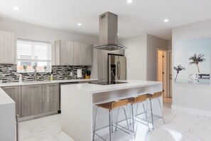 A modern kitchen with a central island, bar stools, and a large window with blinds. - Heated Pool & BBQ Olas Villa by Rovetravel (Fort Lauderdale)