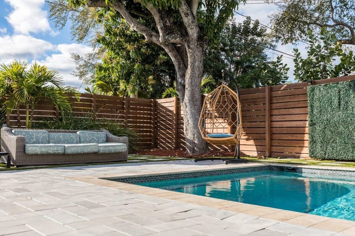 A swimming pool with a wooden fence, a wicker sofa, and a hanging chair.