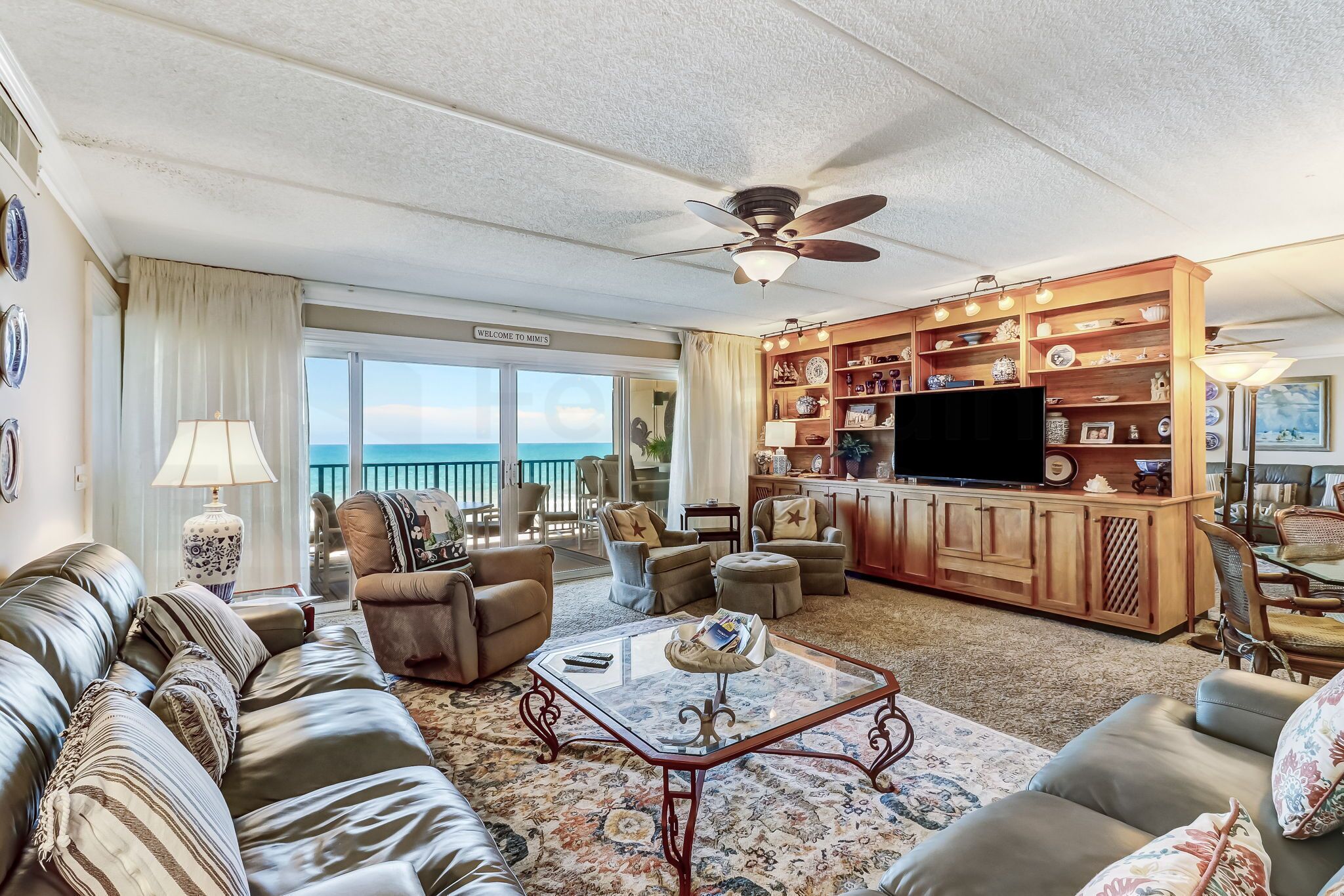 A spacious living room with a large sofa, armchairs, a coffee table, and a TV. There is a wooden cabinet with decorative items and a view of the ocean through large windows.
