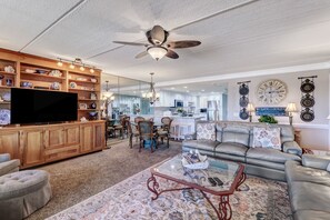 Ein geräumiges Wohnzimmer mit großem Fernseher, Holzregalen, Deckenventilator und Essbereich mit Stühlen und Tisch.