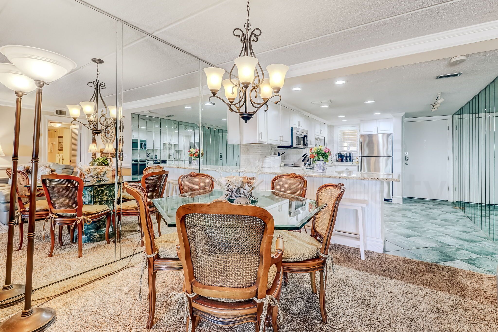 Un comedor con una mesa de vidrio rodeada de sillas de madera, un candelabro y vistas a una cocina moderna.
