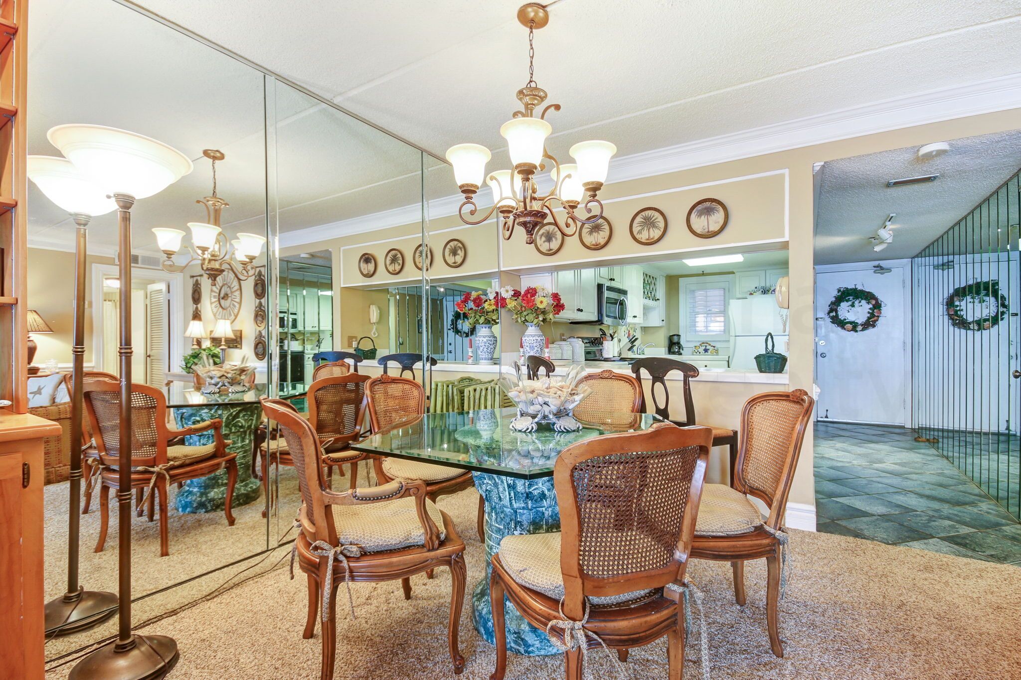 A dining area with a glass table surrounded by wicker chairs, a chandelier, and a mirror reflecting the room.