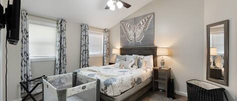 A bedroom with a bed, a ceiling fan, a TV, a crib, a mirror, and a window with curtains.