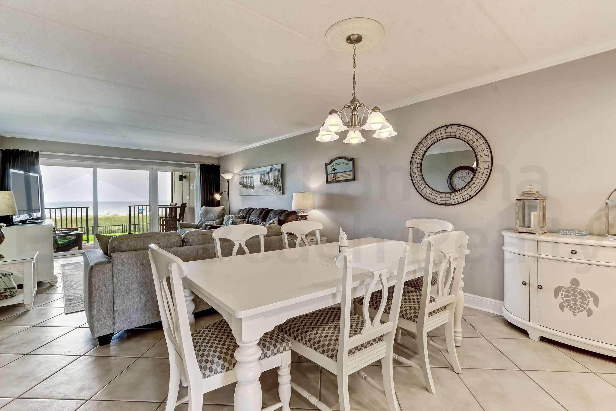 A spacious living area with a dining table, white chairs, a sofa, and a large window offering a view of the outdoors.