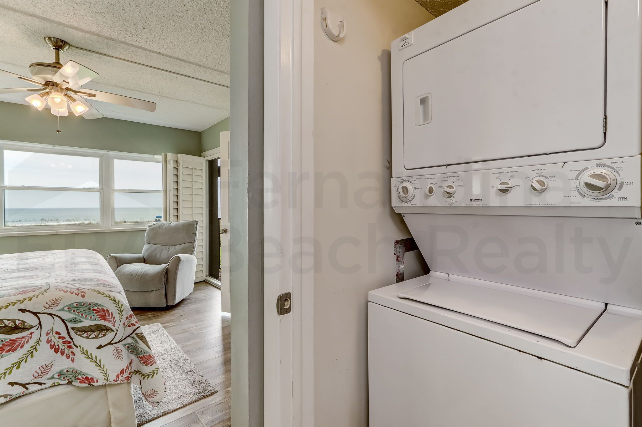 A laundry room with a front-loading washer and dryer, a ceiling fan, and a view through an open door to a bedroom with a bed and a chair.