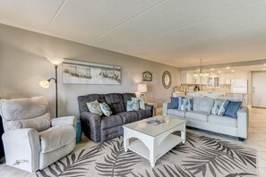 Een woonkamer met een grijze bank, een witte fauteuil en een glazen salontafel. Aan de muur hangt een schilderij en erboven de bank een klok.
