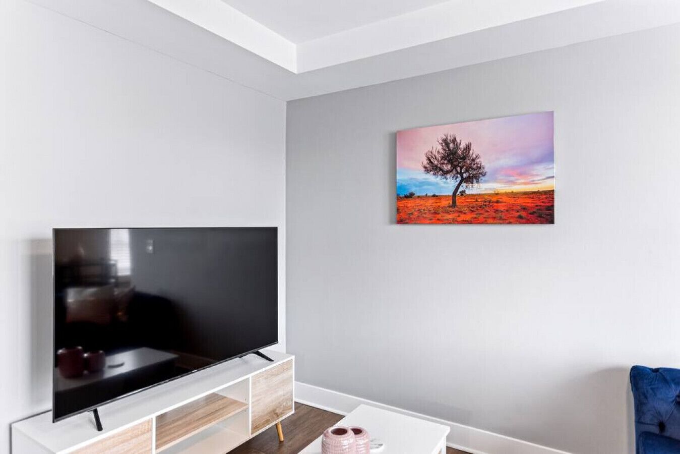 A modern living room with a flat-screen TV on a wooden TV stand, a painting of a tree on a red field, and a blue chair.