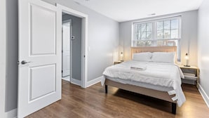 Een slaapkamer met een bed, nachtkastjes, een raam met zonwering en een deur naar een andere kamer.