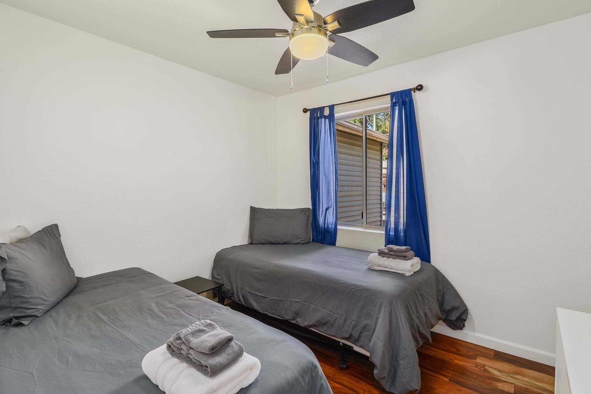 Un dormitorio con dos camas, ventilador de techo y ventana con cortinas azules.