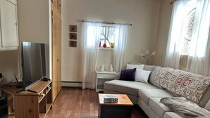 Een woonkamer met een houten vloer, een grijze bank, een salontafel, een televisie op een houten dressoir en een raam met transparante gordijnen.