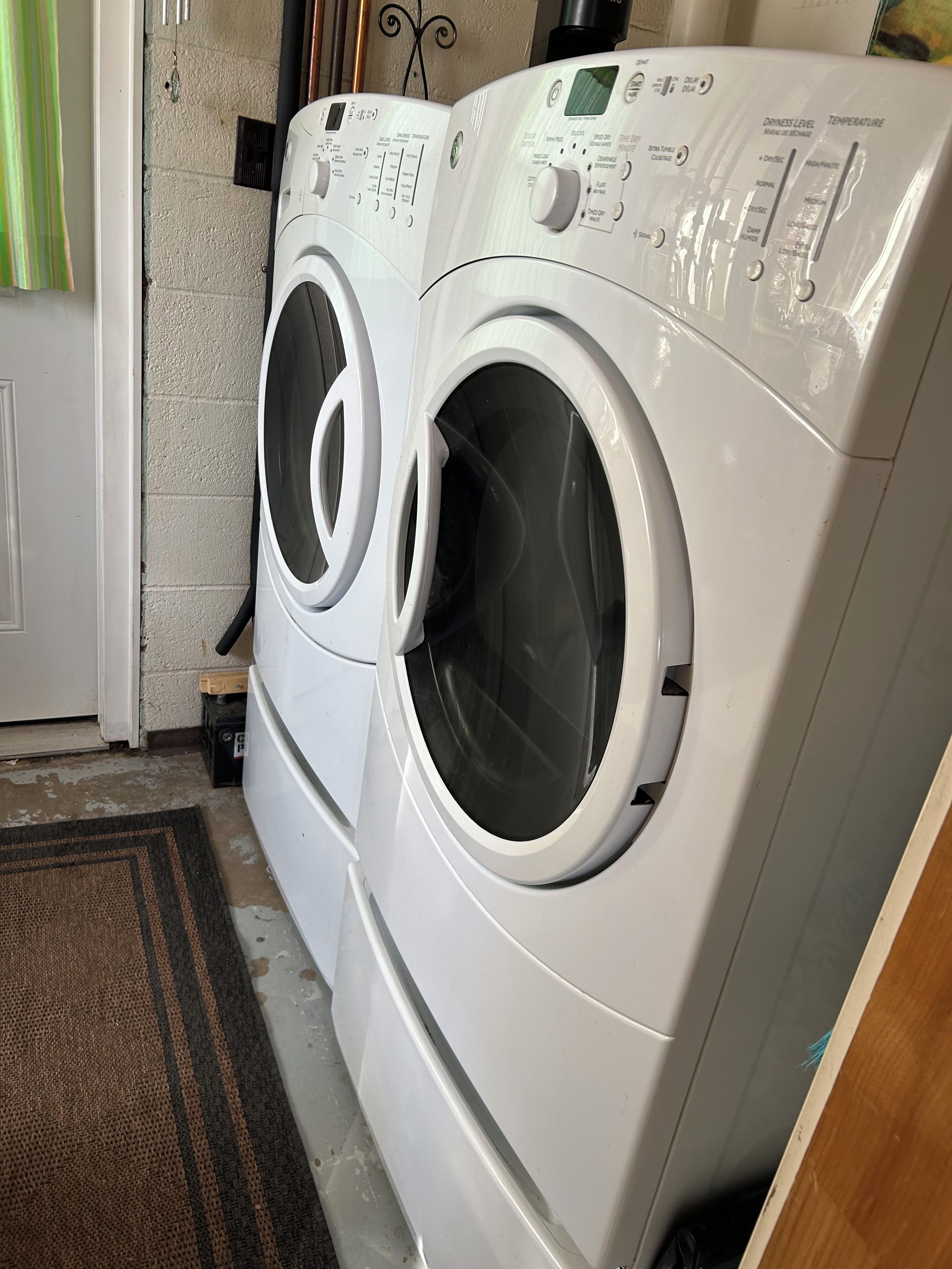 A front-loading washer and dryer unit with control panels, positioned against a wall with a door and a doormat.