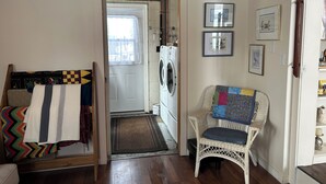 A laundry room with a washing machine, a wicker chair with a quilt, and a door leading to another room. - Cozy cottage in Three Hills, Alberta. Pet Friendly! (Three Hills)