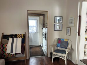 Een wasserette met een wasmachine, een rieten stoel met een sprei, en een deur naar een andere kamer.