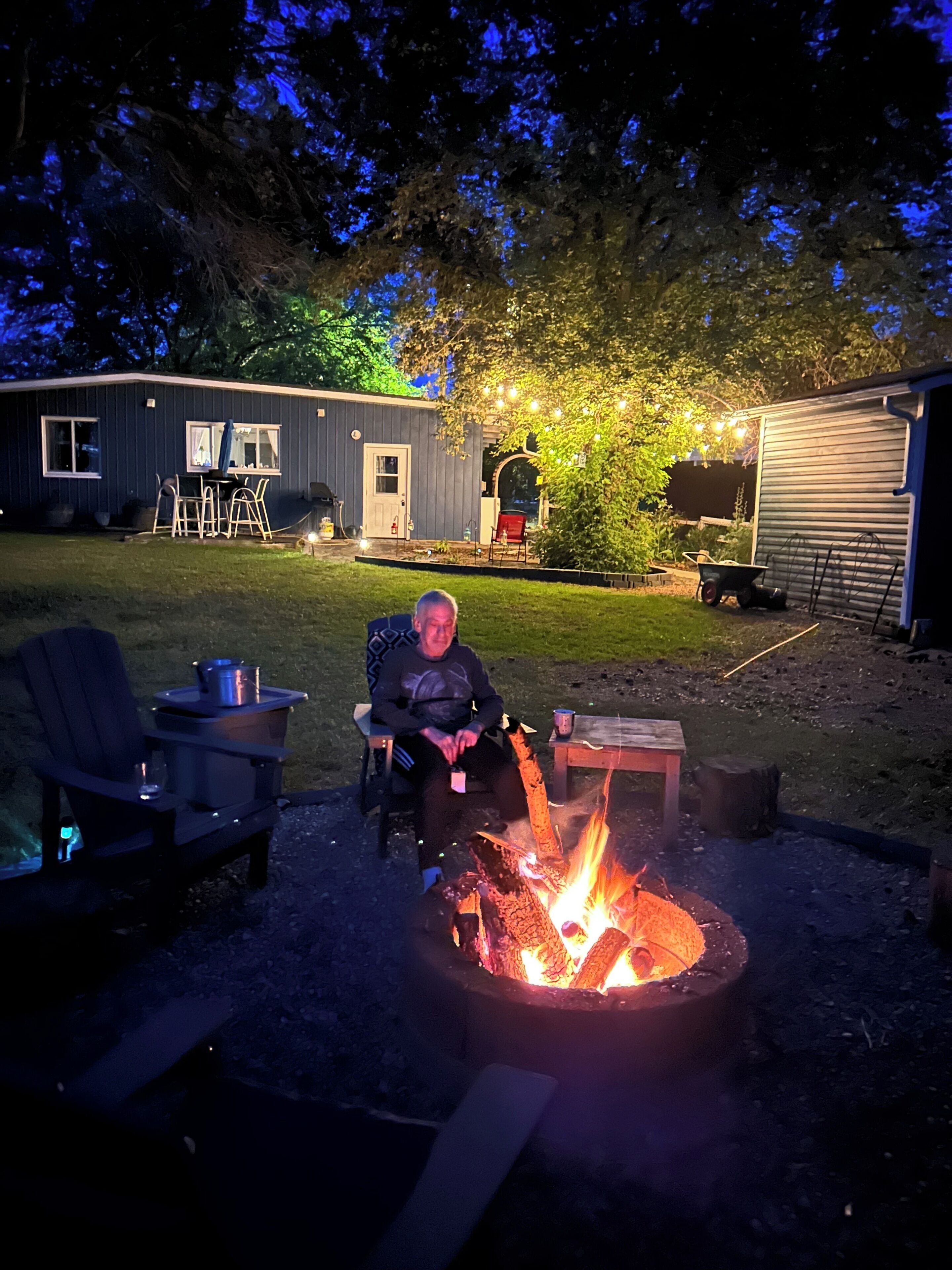 Une personne assise près d’un feu de camp dans un jardin, avec une maison, un abri de jardin et du mobilier d’extérieur.