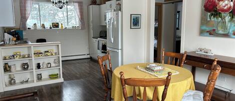 A cozy dining area with a wooden table and chairs, a kitchen in the background, and a painting on the wall.