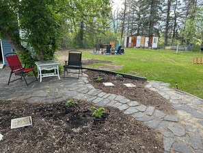 A patio area with stone flooring, a red chair, a white table, and a small garden bed with plants. - Cozy cottage in Three Hills, Alberta. Pet Friendly! (Three Hills)