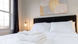 Un letto rifatto con lenzuola bianche e un asciugamano piegato sul lato, una lampada fissata al muro e una finestra con tende.