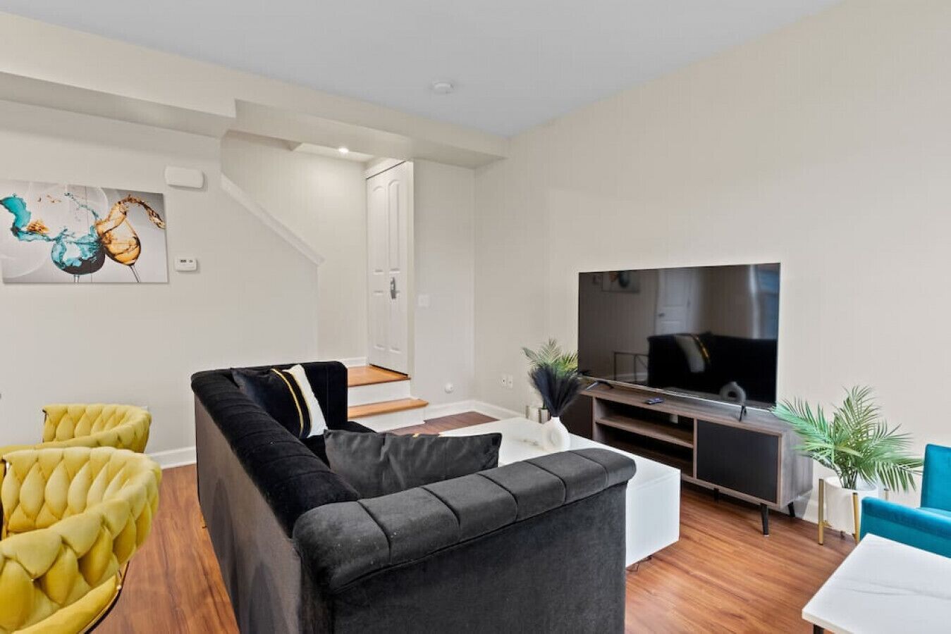 Un salon moderne avec un canapé d’angle noir, des fauteuils jaunes, un téléviseur à écran plat et un escalier menant à un autre niveau.