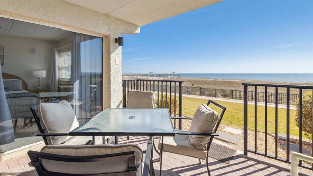 A balcony with a glass door leading to a bedroom, a table with chairs, and a view of the beach. - J1 Amelia South (Fernandina Beach)