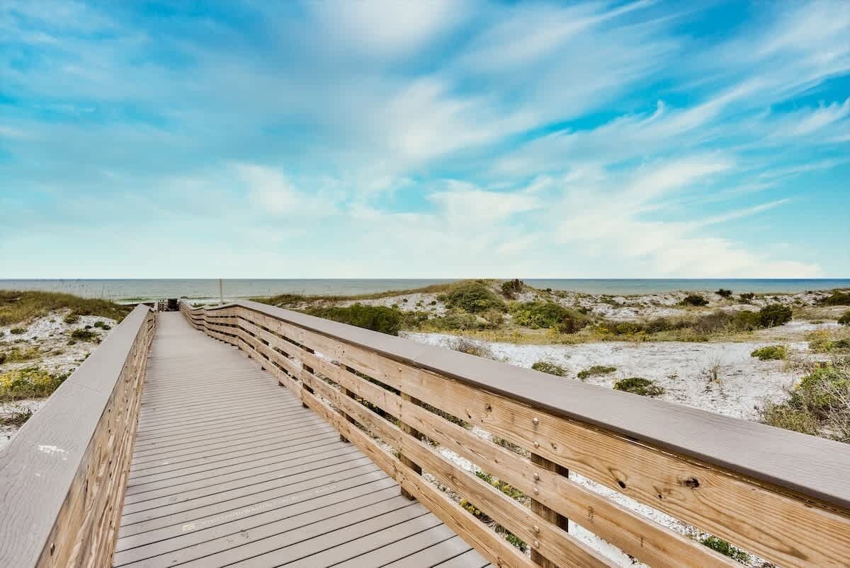 A wooden boardwalk leading to a beach with dunes and a clear sky.