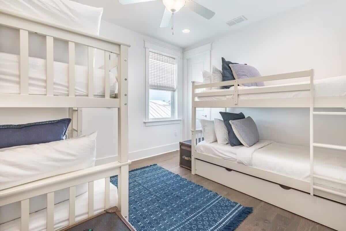 A room with bunk beds, a window with blinds, a ceiling fan, and a blue rug.