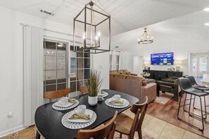 Ein moderner Essbereich mit einem schwarzen Tisch, gedeckt für vier Personen, umgeben von Holzsesseln. Ein großes Fenster mit Jalousien ist sichtbar, und ein Fernseher ist an der Wand montiert.