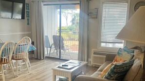 Ein Wohnzimmer mit Sofa, Couchtisch und Essbereich mit Stühlen. Eine Glastür führt zu einem Außenbereich mit Blick auf Bäume und einem Geländer.