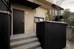 A residential building with a dark wooden door, beige walls, and a small set of steps leading to a balcony. - Nozomu House02 (Kyoto)