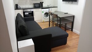 A compact living space with a sofa, a small table, a washing machine, and a microwave in the kitchen area. - Cosy Central Cardiff Retreat (Cardiff)
