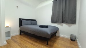A bedroom with a bed, a nightstand, a lamp, a curtain, and a trash bin. - Cosy Central Cardiff Retreat (Cardiff)