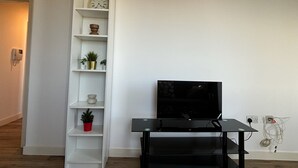 A modern living room with a black TV stand, white shelving unit with decorative items, and a clock. - 4 Bed 2 Bath Balcony Retreat - 15min-O2 Arena! (London)