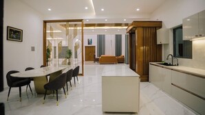 Una cocina moderna con una isla central, una zona de comedor con una mesa larga y sillas, y una sala de estar con un sofá y cortinas.