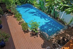Una piscina rectangular con una terraza de madera alrededor, plantas en macetas y vista al cielo.