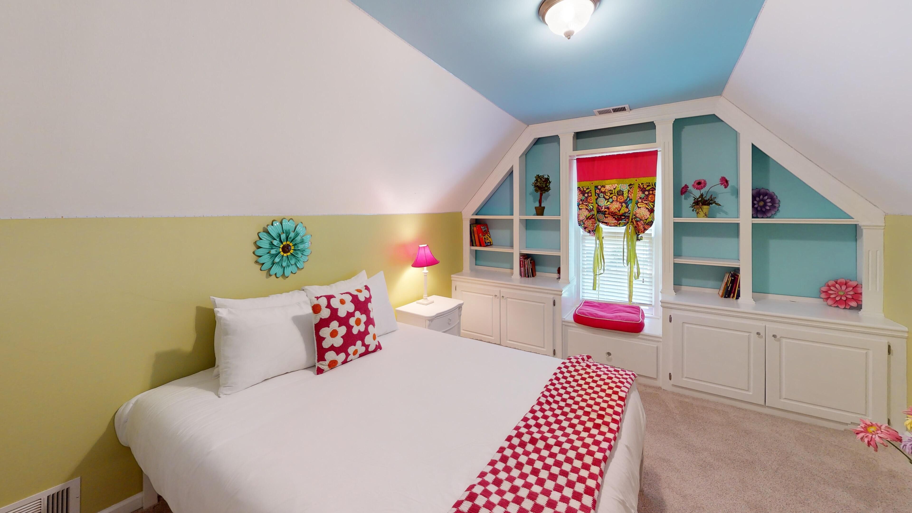 A bedroom with a bed, a nightstand, a lamp, a window with blinds, and a built-in shelving unit with books and decorative items.